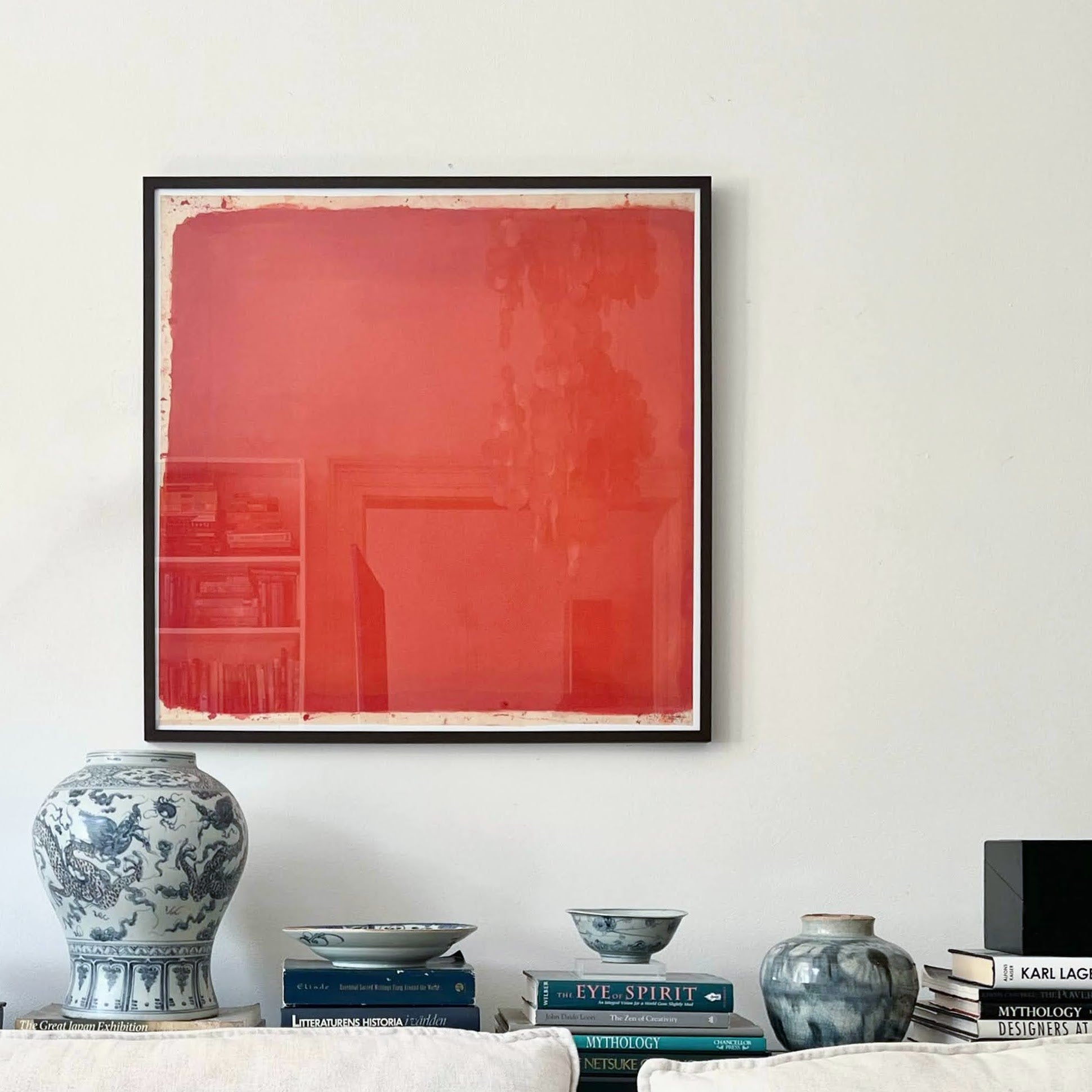 Framed abstract red painting on a wall above a shelf with decorative items.