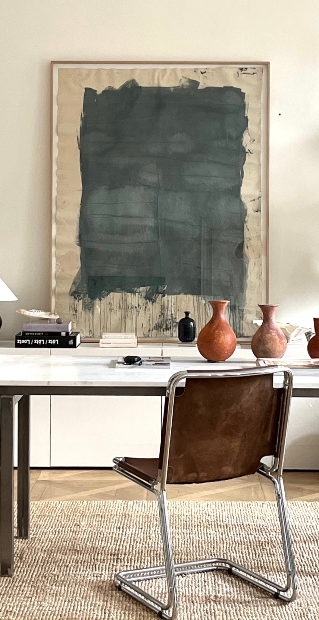 A large abstract painting in shades of green and blue, mounted on a wall above a white desk and a chair with a wooden frame, with decorative objects and a vase on the desk.