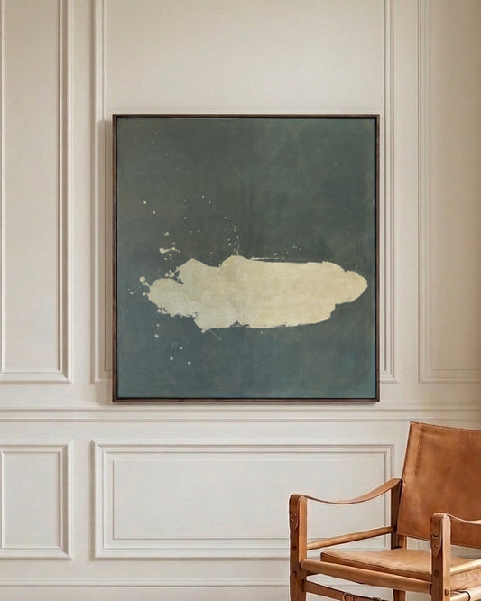A square abstract wall art print with a dark slate blue background and a textured cream shape in the center, displayed on a cream paneled wall above a mid-century modern wooden armchair.