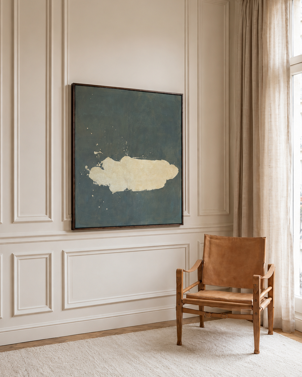 A framed abstract painting with a dark slate blue background and a textured cream cloud-like shape, displayed on a paneled cream wall in a bright room next to a wooden armchair and a window with beige curtains.