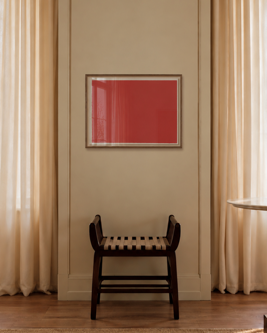 A minimalist interior scene featuring a small dark wooden chair with a woven seat, centered against a beige wall beneath a framed red square artwork. The room has sheer cream curtains, tall windows with bare tree branches outside, a light wood floor, and a textured beige rug.