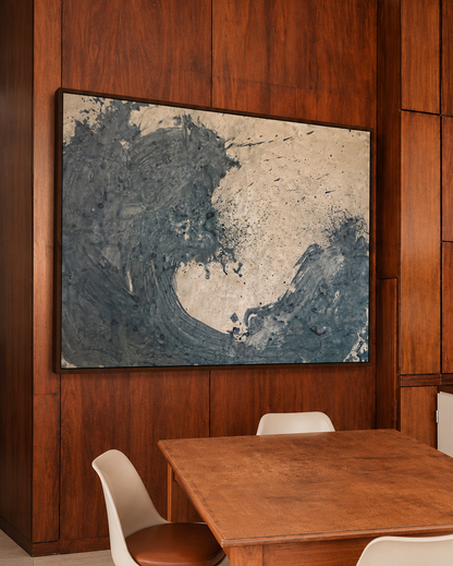 A large abstract wave painting in blue, gray by Nicolas Makelberge, and beige hangs on a dark wood-paneled wall above a wooden dining table with white chairs in a modern dining room.