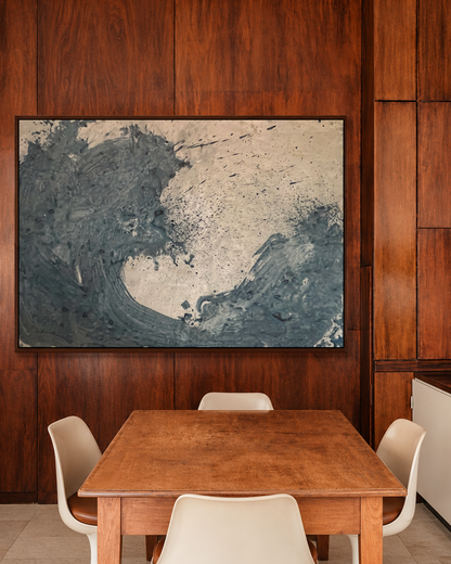 A large abstract wall art piece with swirling blue-gray and cream tones by Nicolas Makelberge hangs on a dark wood-paneled wall above a wooden dining table with white chairs in a modern interior setting.