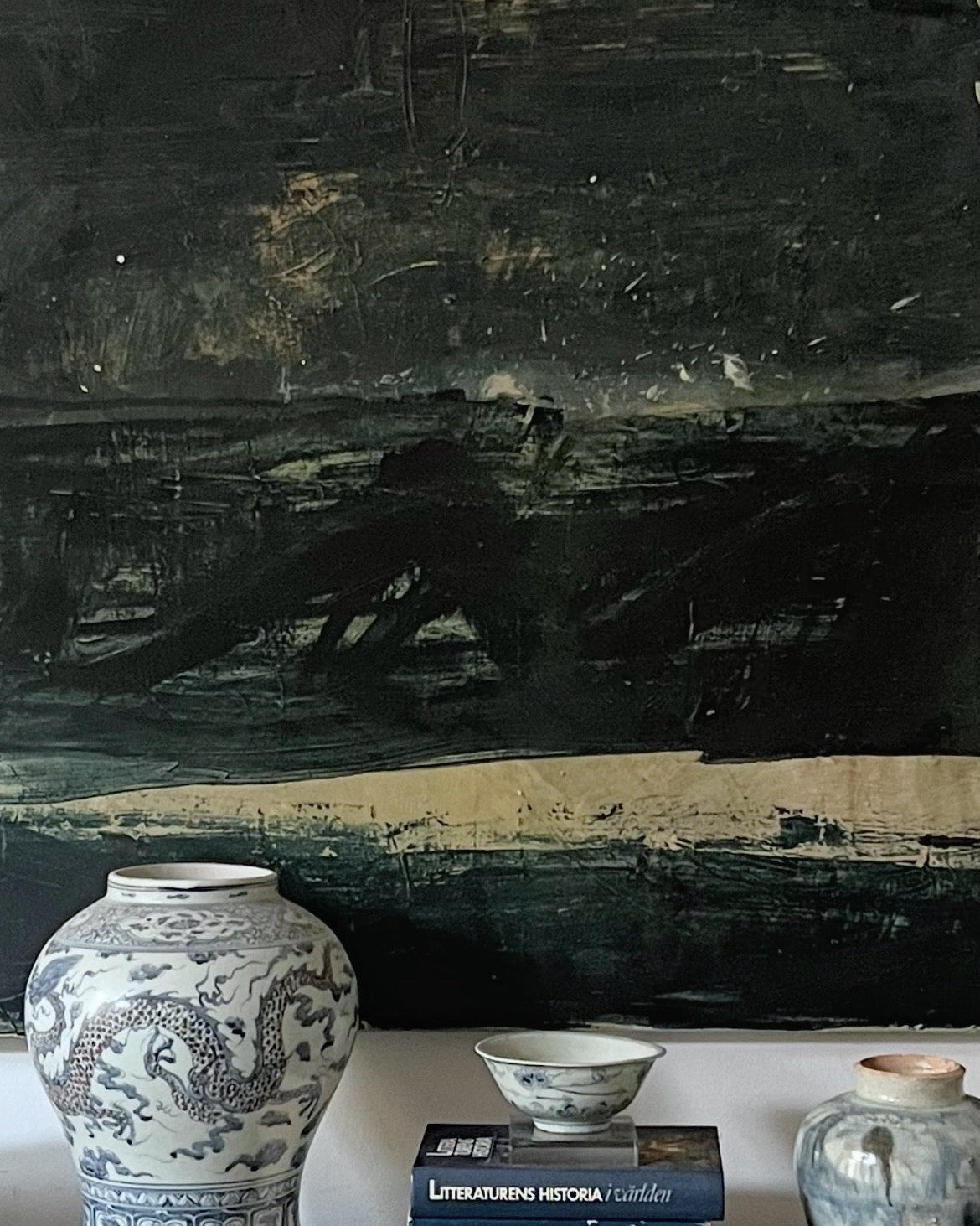 A large black abstract wall art piece hangs above a console table holding a blue-and-white porcelain vase, a small bowl, books, and another ceramic vessel in a living room setting.