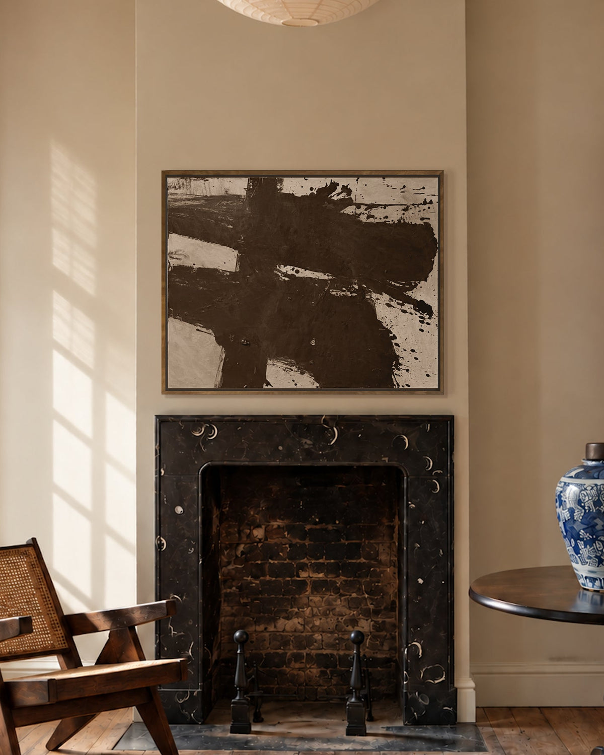 A styled interior room with a black marble fireplace, a large abstract painting above it by Nicolas Makelberge, a wooden armchair by a window, a round black side table with a blue-and-white ceramic vase, and a woven rug on a wood floor.