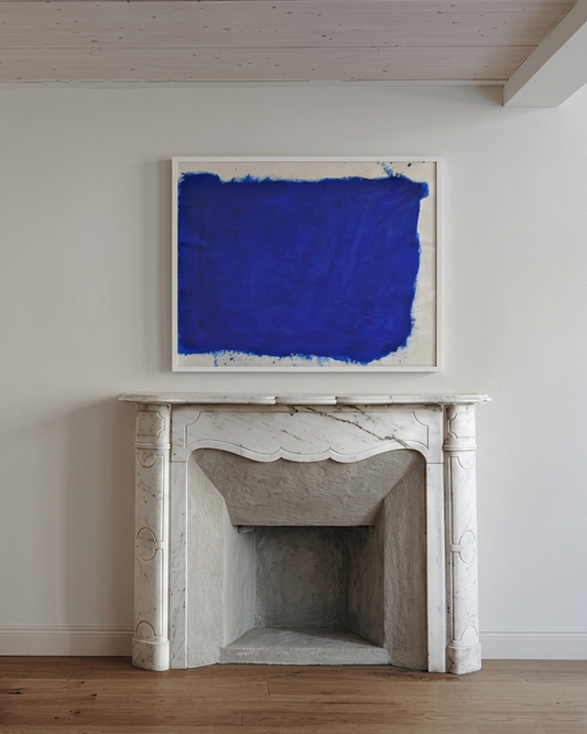 Blue abstract painting above a marble fireplace in a room with wooden flooring.