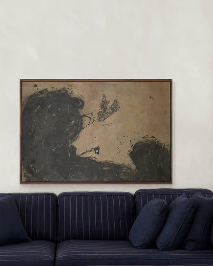 An abstract framed painting with dark charcoal and beige tones hangs on a white wall above a navy blue pinstriped sofa in a modern living room setting.