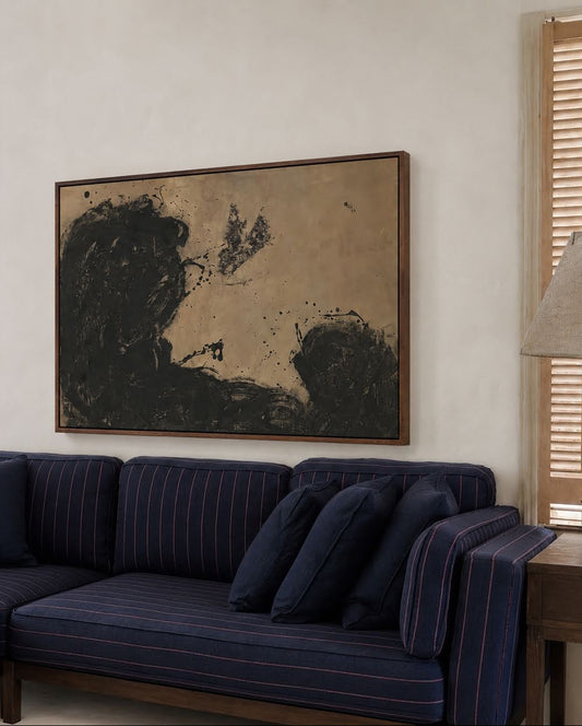 A large framed abstract painting with black and dark brown brushstrokes on a warm beige background, displayed on a cream wall above a navy blue sectional sofa in a styled living room.
