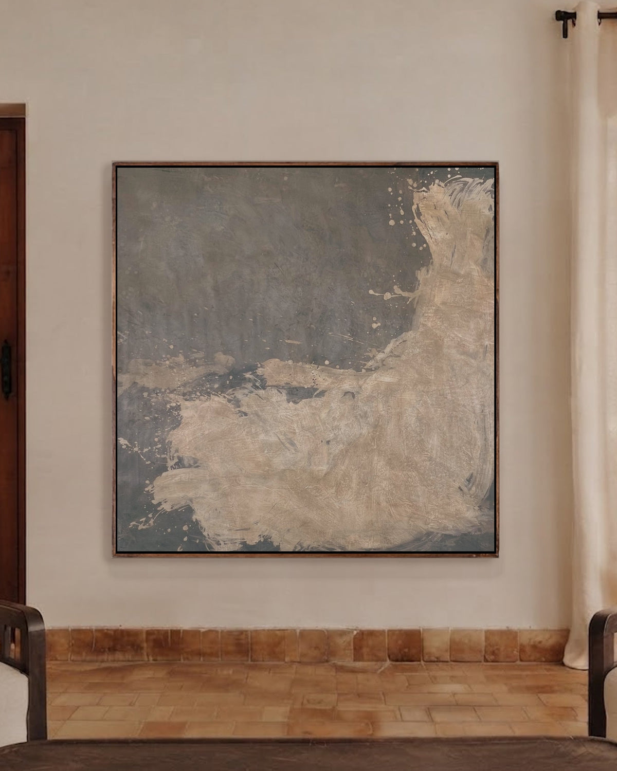 A large square abstract canvas print with textured gray, slate blue, and beige tones, displayed on a warm beige wall in a modern living room setting with two cream armchairs and a dark wooden coffee table.
