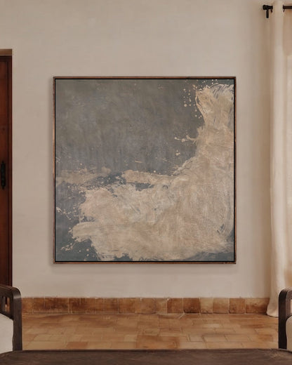 A large square abstract canvas print with textured gray, slate blue, and beige tones, displayed on a warm beige wall in a modern living room setting with two cream armchairs and a dark wooden coffee table.
